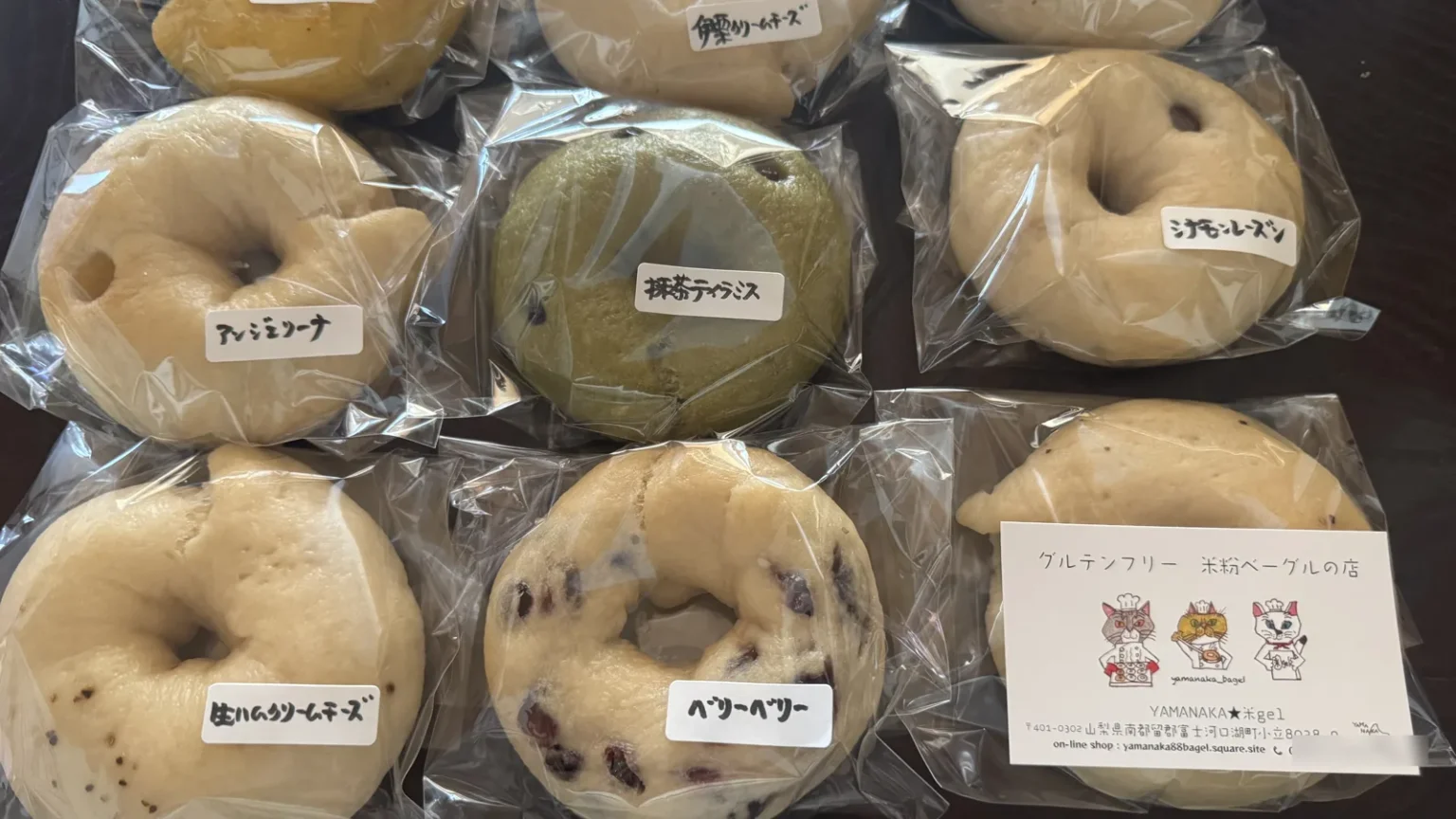 Bagged rice-flour bagels with handwritten Japanese flavour labels and a shop card reading グルテンフリー 米粉ベーグルの店