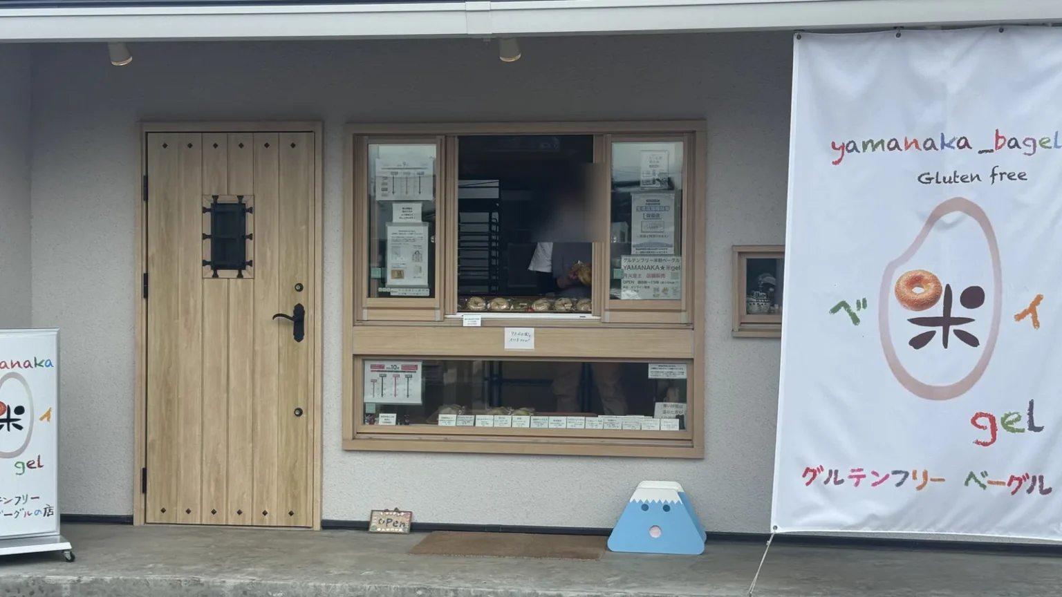 A small shopfront with a wooden door, a serving window with notices and a white banner reading yamanaka_bagel Gluten free