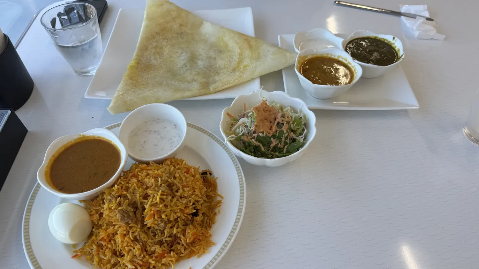 A plate of biryani with a boiled egg, a dosa on a square plate, small bowls of curry, a small white dip and a salad, on a white table