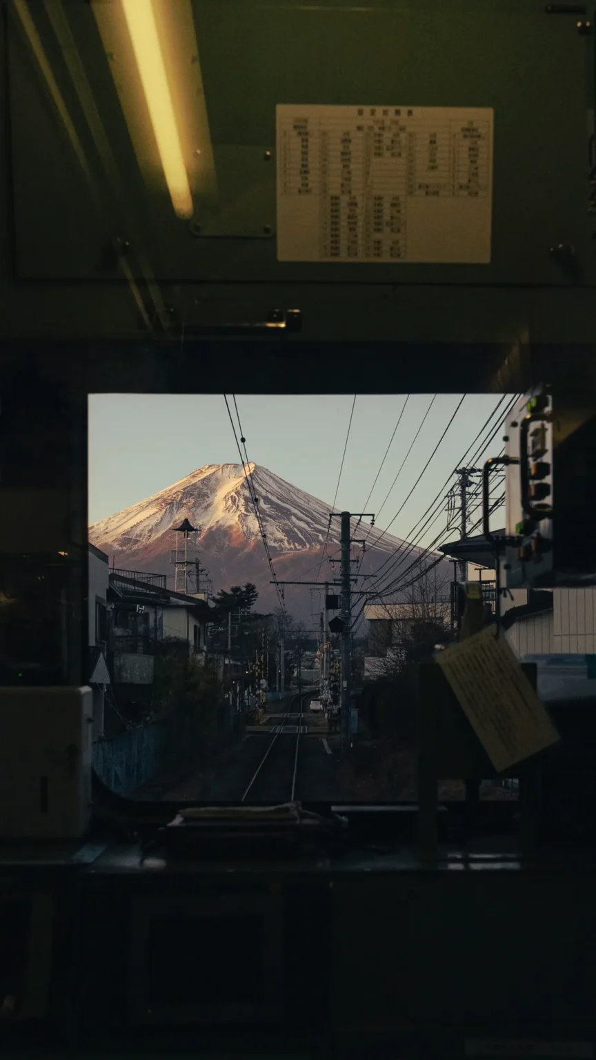 View from behind a train driver's cab: a posted timetable sheet above, and through the front window a single track running ahead towards Mt Fuji, its snowy summit lit pink.