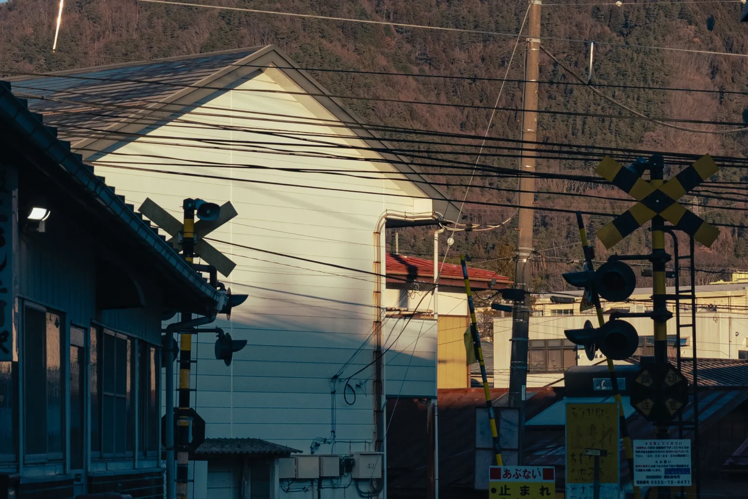 Railway crossing signals with yellow-and-black crossbucks and raised barrier arms beside buildings in Fujiyoshida, below a wooded hillside lit by the morning sun.
