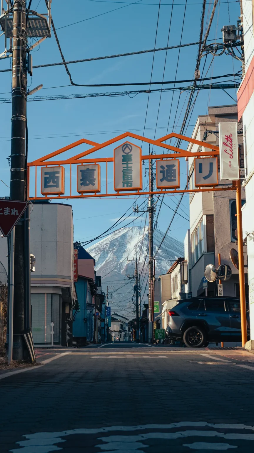 The orange Nishiura-dori arch reading 西裏通り over a street of low shops and power lines in Fujiyoshida, with a car parked on the right and the snowy cone of Mt Fuji at the far end.