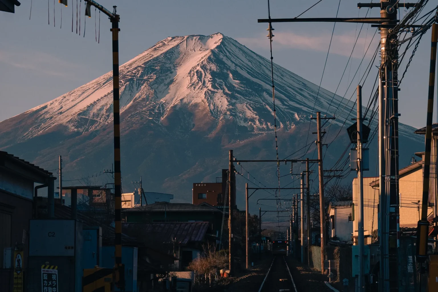 A single railway track with overhead wires runs between low buildings towards Mt Fuji, its snowy upper slopes tinted pink by the early sun.
