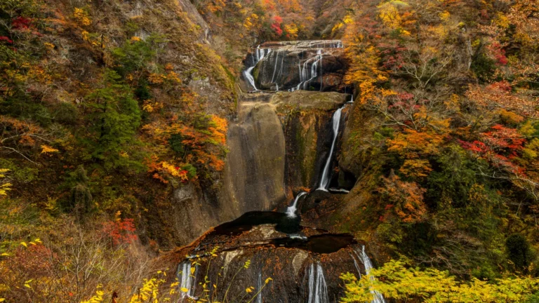 A tiered waterfall dropping over layered grey rock between slopes of orange, red and yellow autumn trees