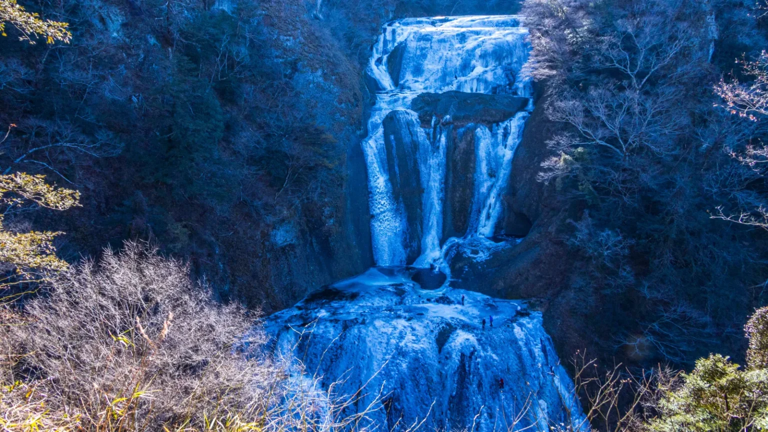 A waterfall partly covered in white and blue ice, with bare trees and frosted slopes around it