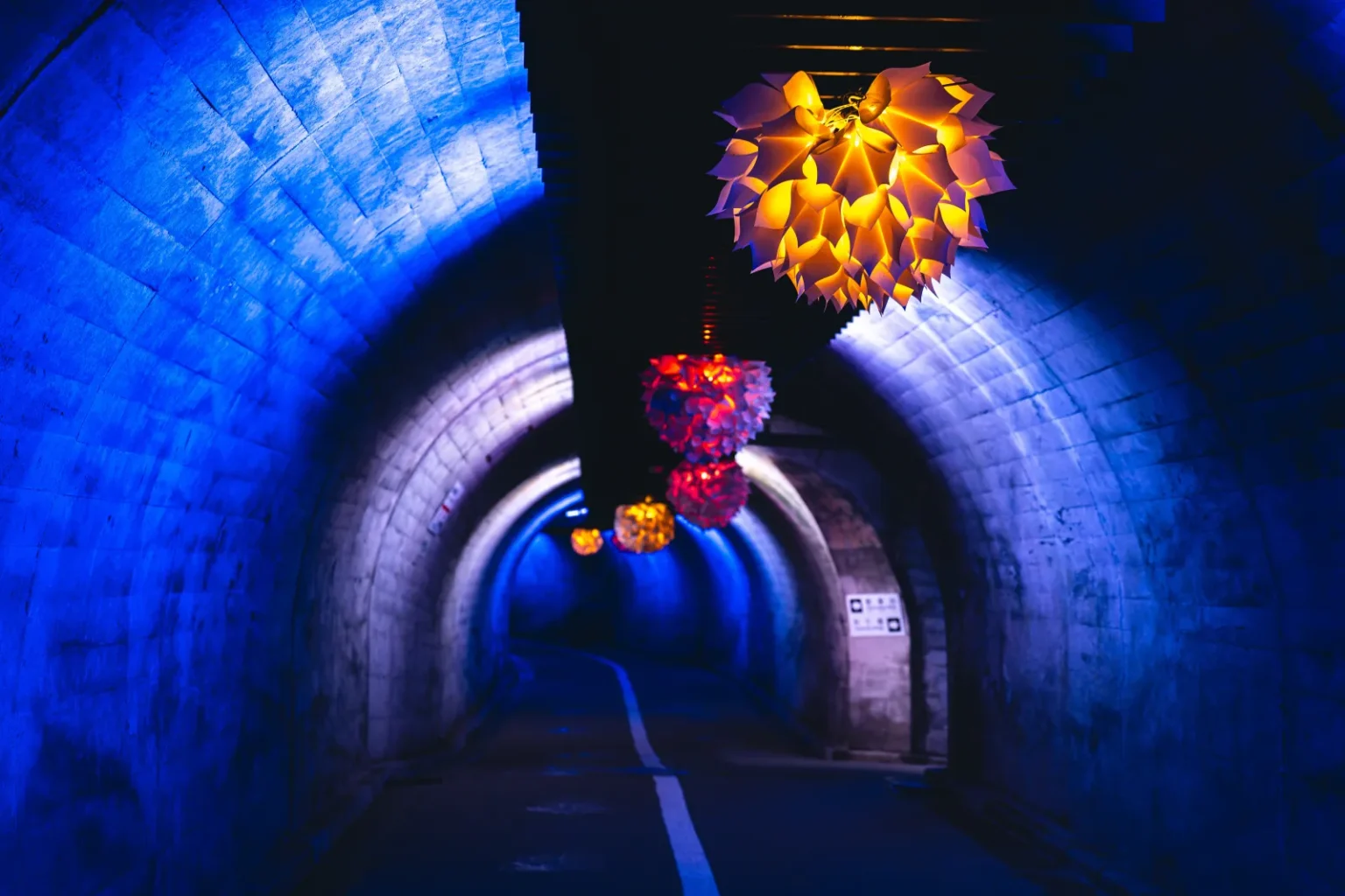 A long arched tunnel lit in blue, with glowing orange and pink paper lanterns hanging from the ceiling