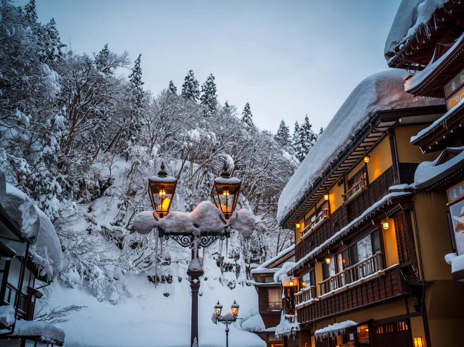 A gas lamp topped with snow beside a snow-covered wooden inn in Ginzan Onsen by day