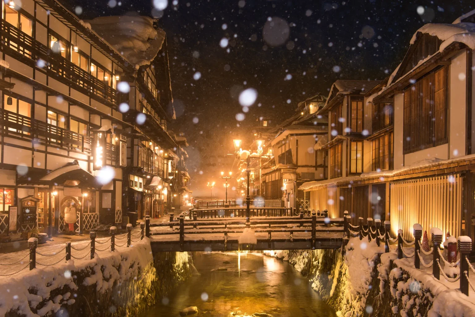 Snow falling on Ginzan Onsen at night, with gas lamps and lit wooden inns on both banks of the river
