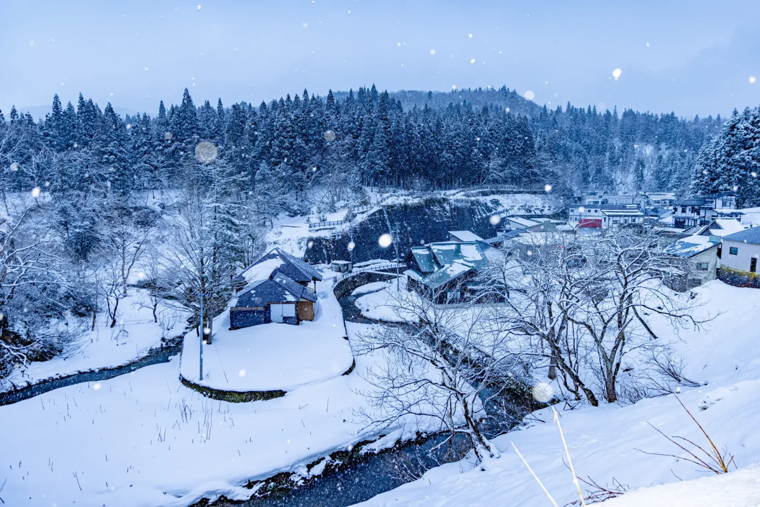 Snow-covered houses, trees and a stream in the Ginzan area of Obanazawa on an overcast winter day