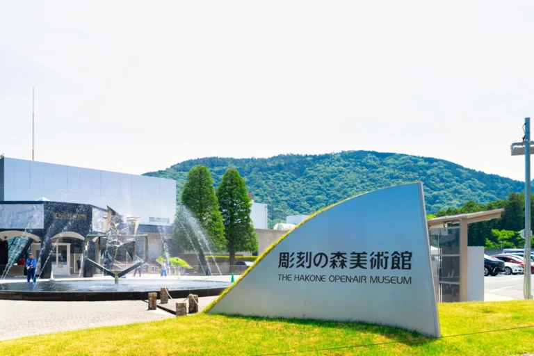 A curved white sign reading 彫刻の森美術館 THE HAKONE OPEN-AIR MUSEUM on a lawn, with a fountain, museum buildings and a wooded hill behind