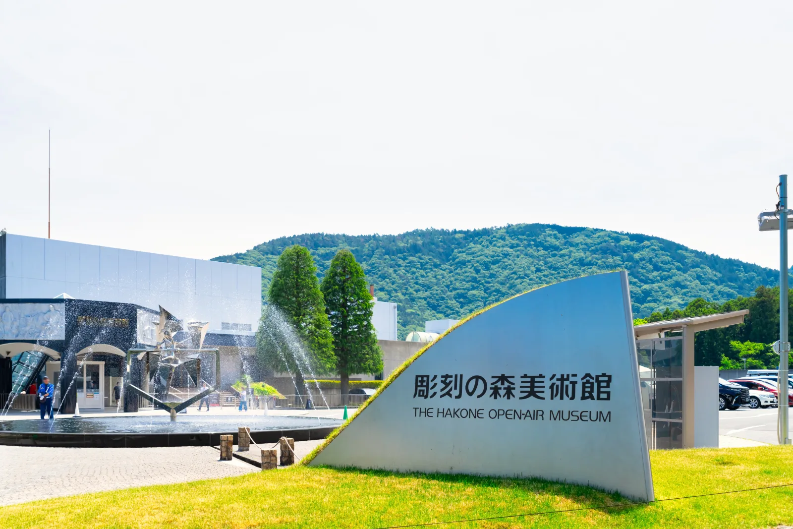 A curved white sign reading 彫刻の森美術館 THE HAKONE OPEN-AIR MUSEUM on a lawn, with a fountain, museum buildings and a wooded hill behind