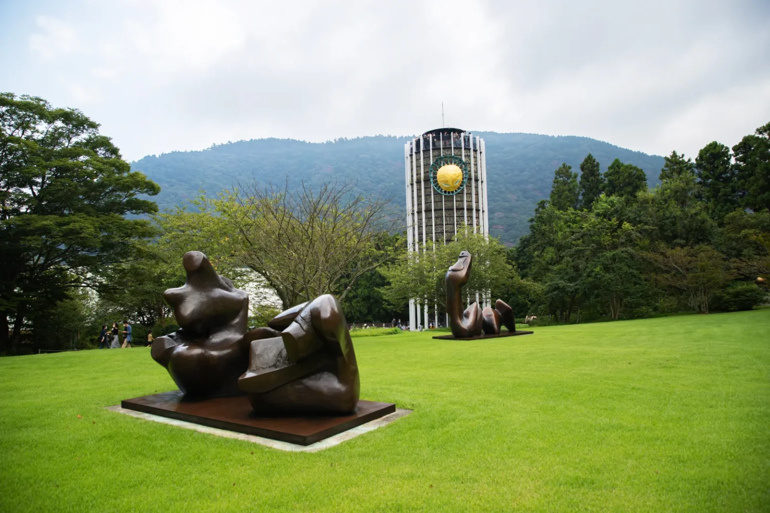 Two large bronze sculptures on a wide green lawn, with a tall white tower, trees and a forested hill behind
