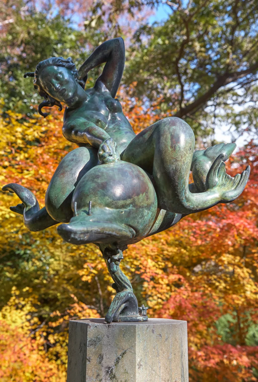 A bronze figure on a winged creature on a stone plinth, against red, orange and yellow autumn leaves