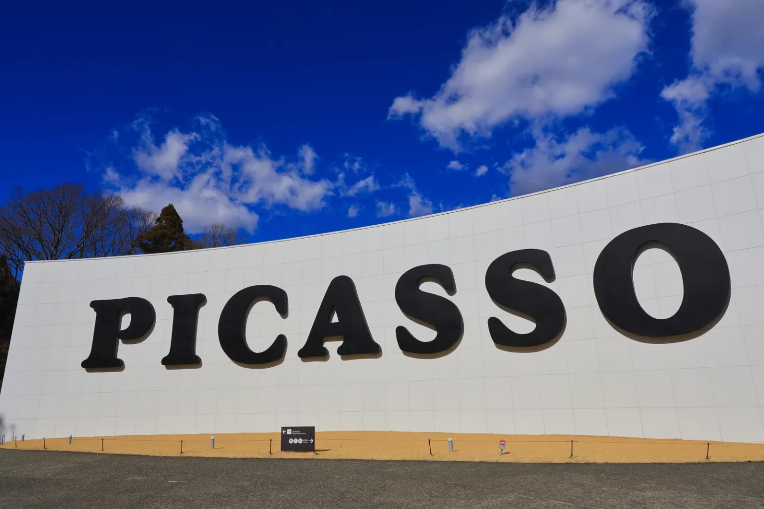 A curved white wall with PICASSO in large black letters under a blue sky with clouds