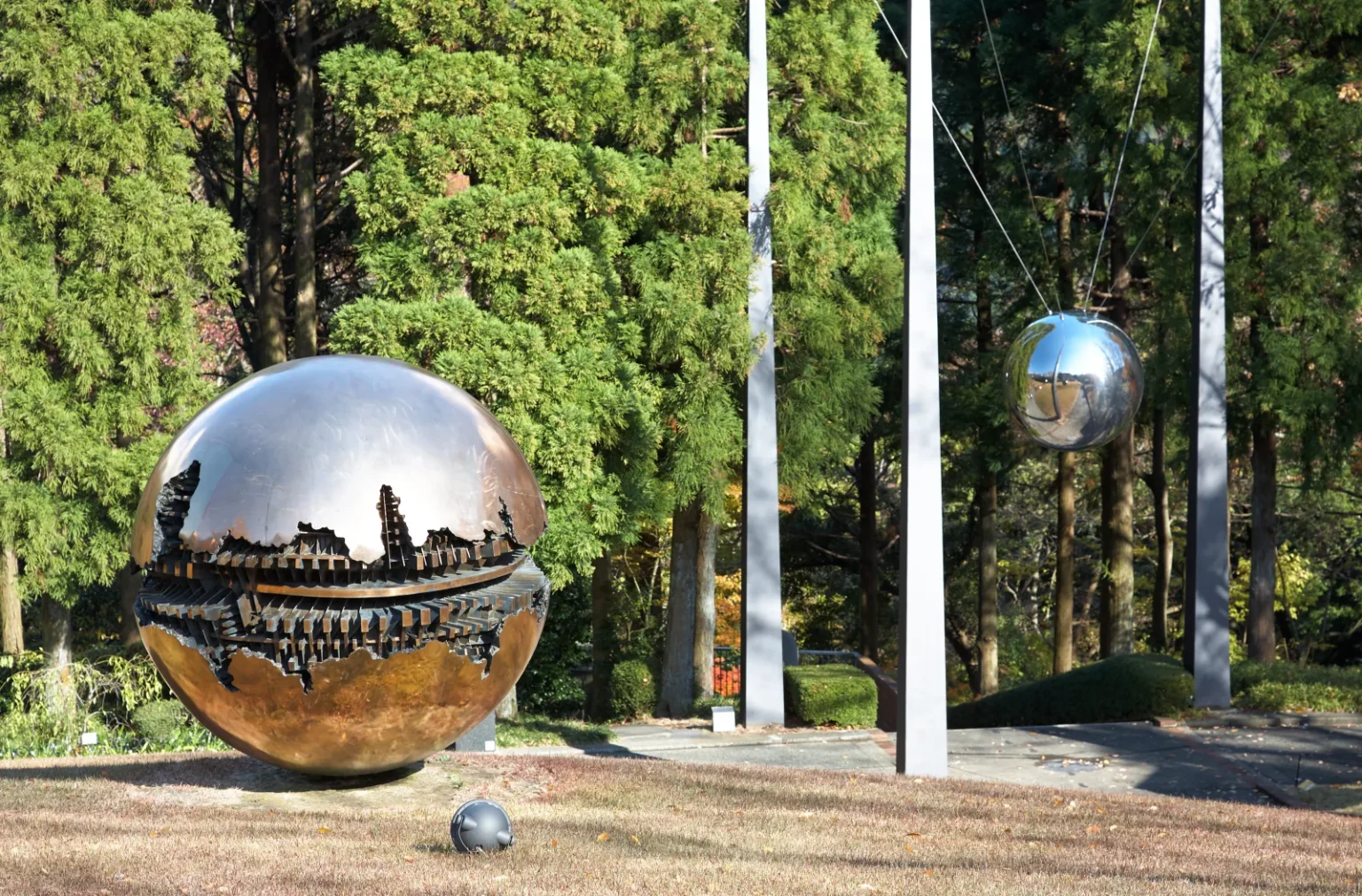 A large bronze sphere with a cracked surface showing inner workings, on gravel in front of tall cedar trees, with a polished steel ball hanging between white poles behind