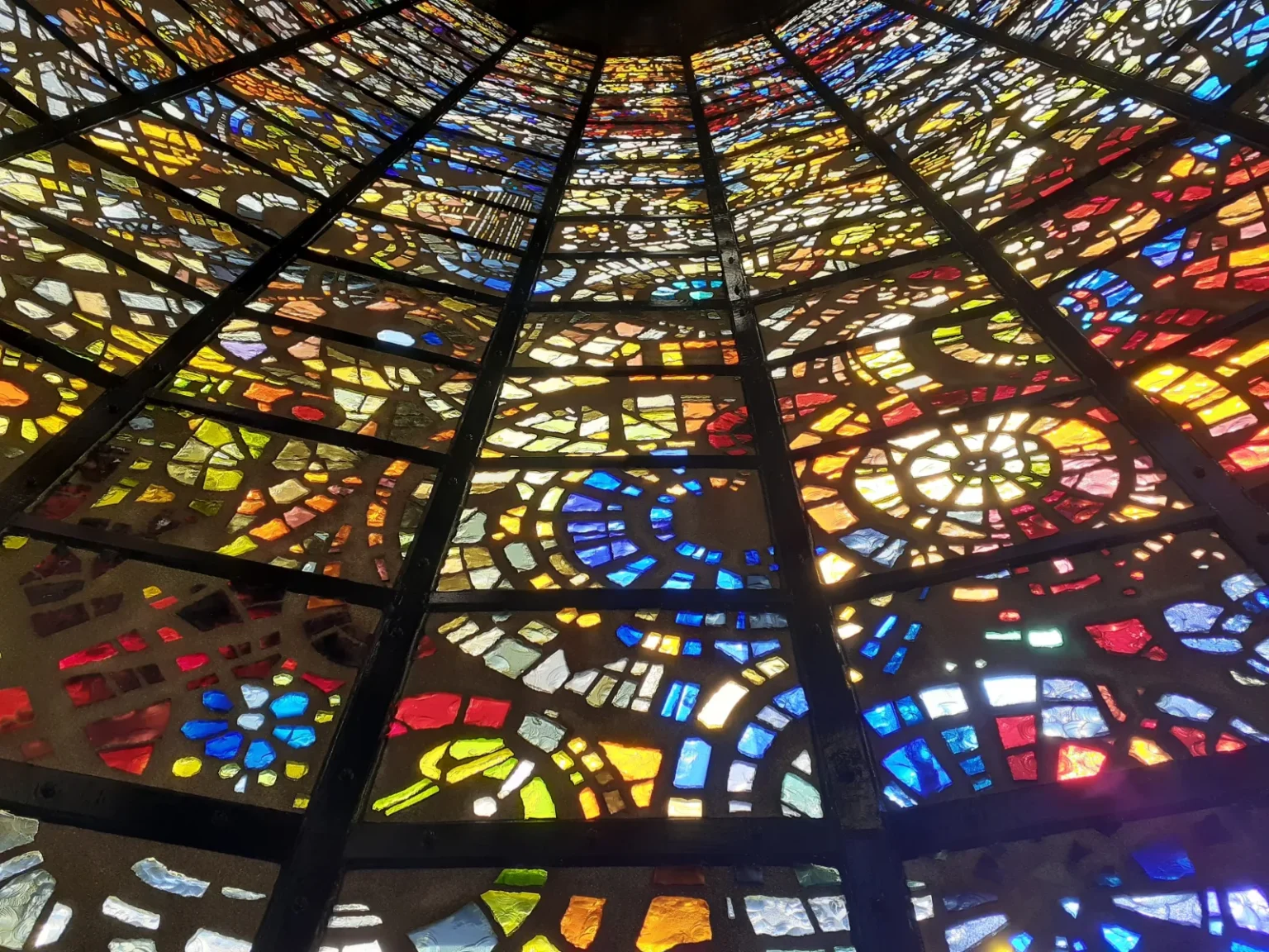 Looking up at a ceiling of stained-glass panels in reds, yellows, blues and greens, divided by dark ribs