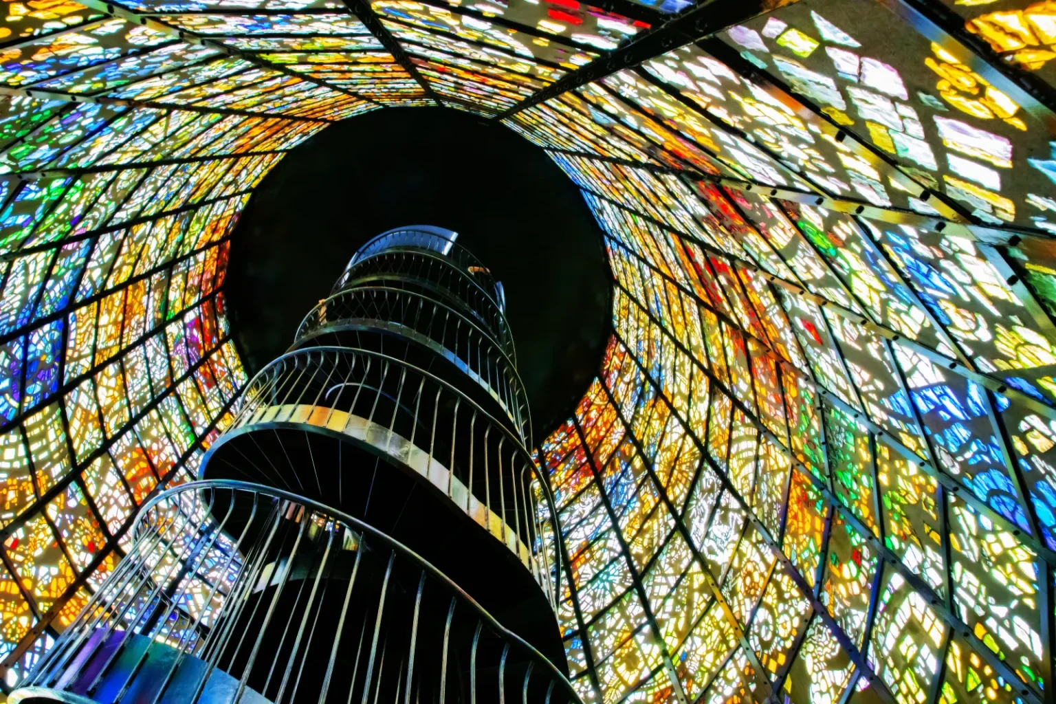 A dark spiral staircase rising through the centre of a round tower lined with colourful stained glass