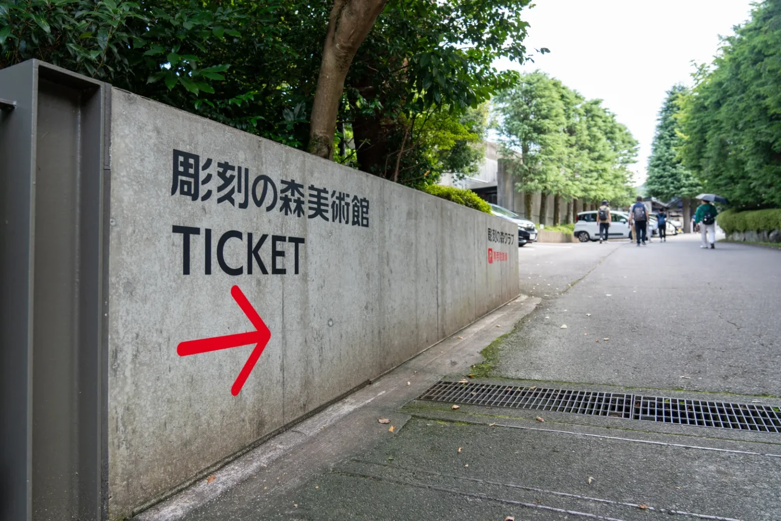 A concrete wall with the sign 彫刻の森美術館 TICKET and a red arrow, beside a sloping path lined with trees