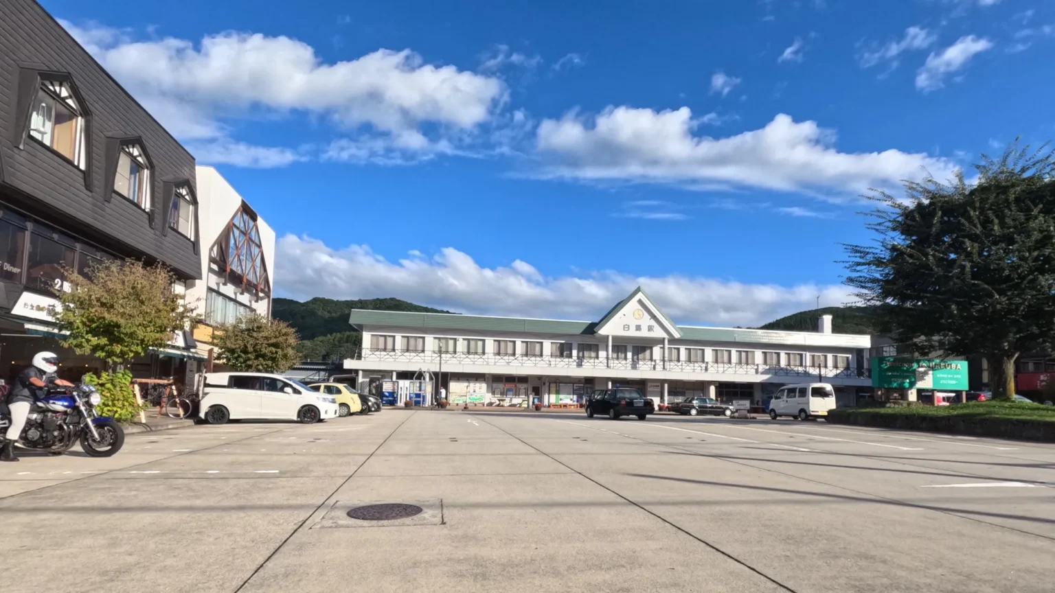 The white two-storey Hakuba Station building with a green roof and the sign 白馬駅, seen across the empty station forecourt under a blue sky with white clouds