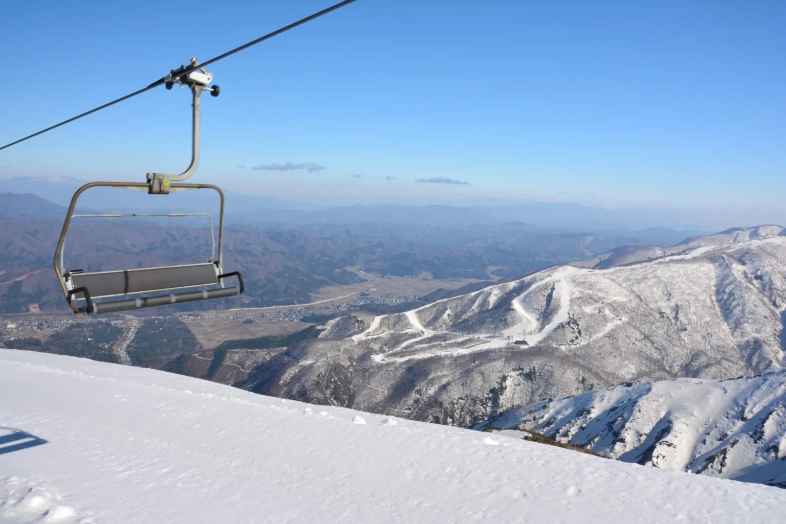 An empty chairlift seat on its cable above a snowy ridge, with the cut runs of Hakuba47 on the mountainside across the valley and the Hakuba basin below
