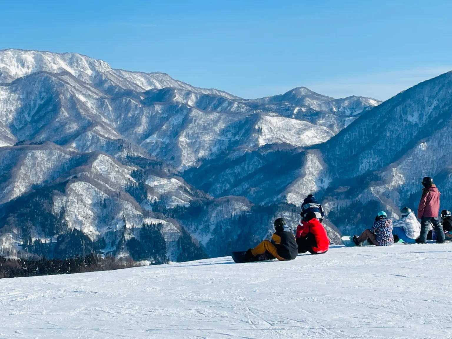 Snowboarders sitting and standing on a groomed slope, taking a break, with forested snowy mountains filling the view behind them
