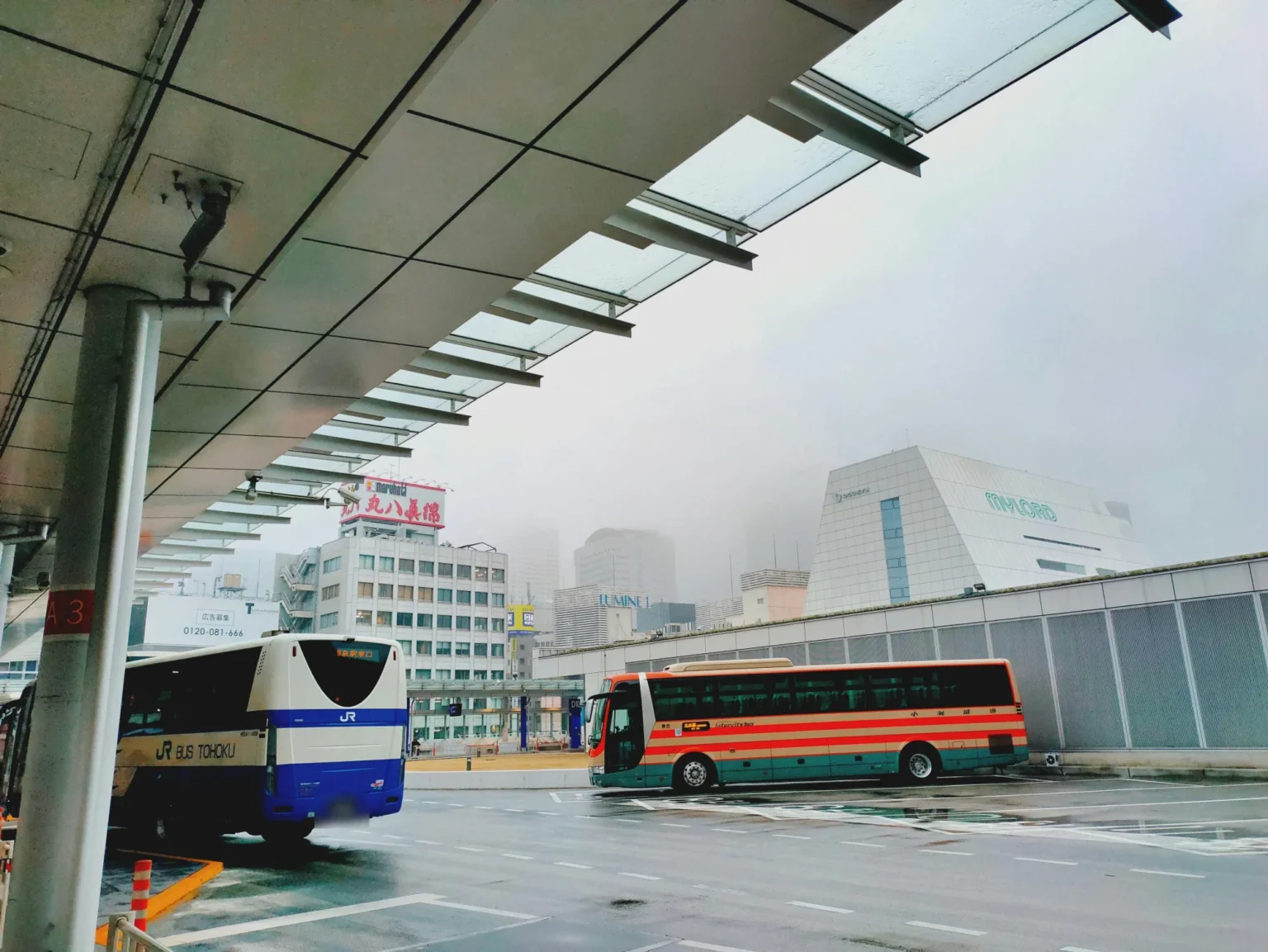 Two highway coaches parked on the open-air bus deck at Busta Shinjuku on a misty, wet day, a blue and white JR Bus Tohoku coach on the left and a red, cream and green striped coach on the right, under a long canopy with Shinjuku buildings behind.