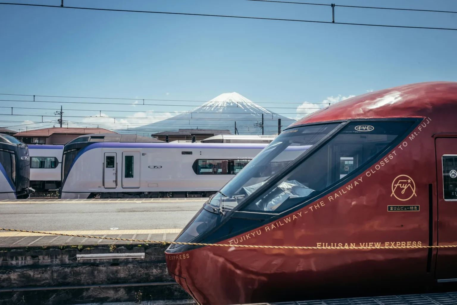 The deep red nose of a Fujikyu Fujisan View Express train lettered Fujikyu Railway, the railway closest to Mt. Fuji, beside white E353-series JR trains with purple stripes, under overhead wires with snow-capped Mt Fuji in a clear blue sky.