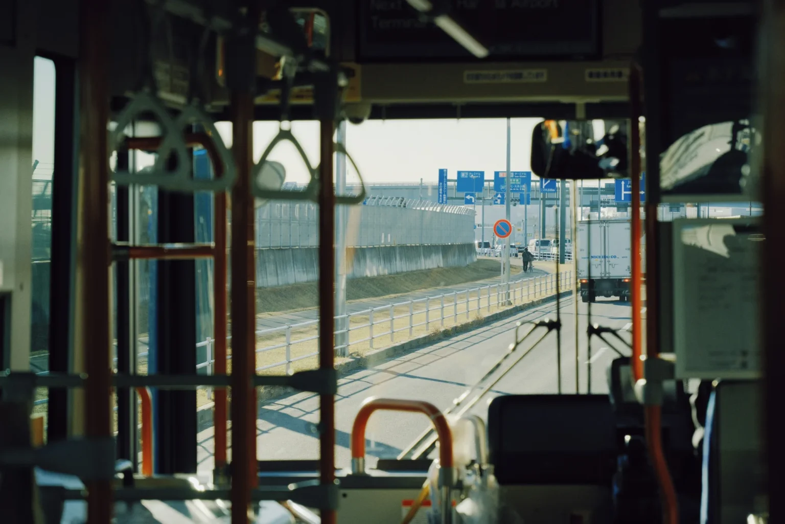 View forward through the windscreen of a bus on an airport access road at Haneda, framed by orange grab poles and hanging straps, with blue direction signs, a security fence and a white delivery truck ahead.