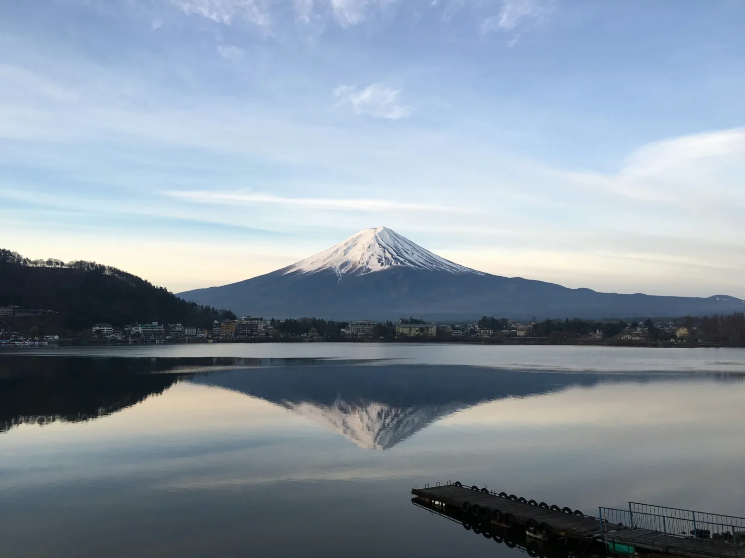Snow-capped Mt Fuji reflected in the still water of Lake Kawaguchi under a pale morning sky, with a wooded ridge on the left, low houses along the far shore and a small pier in the foreground.