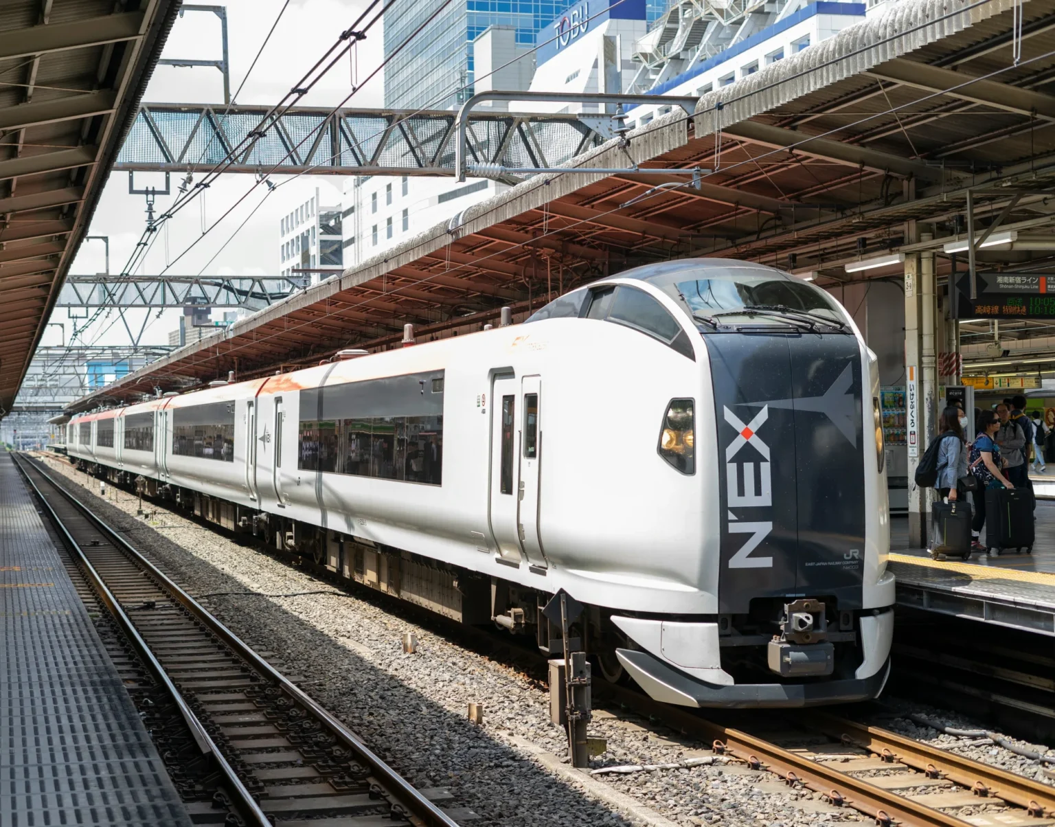 A white, black and red Narita Express E259-series train with the N'EX logo on its black nose standing at an open-air platform under a steel canopy, with tall buildings behind.