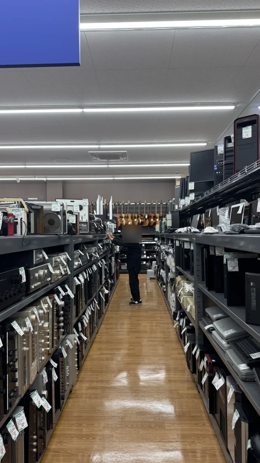 A long aisle of audio equipment, with guitars at the far end and a shopper with a blurred face