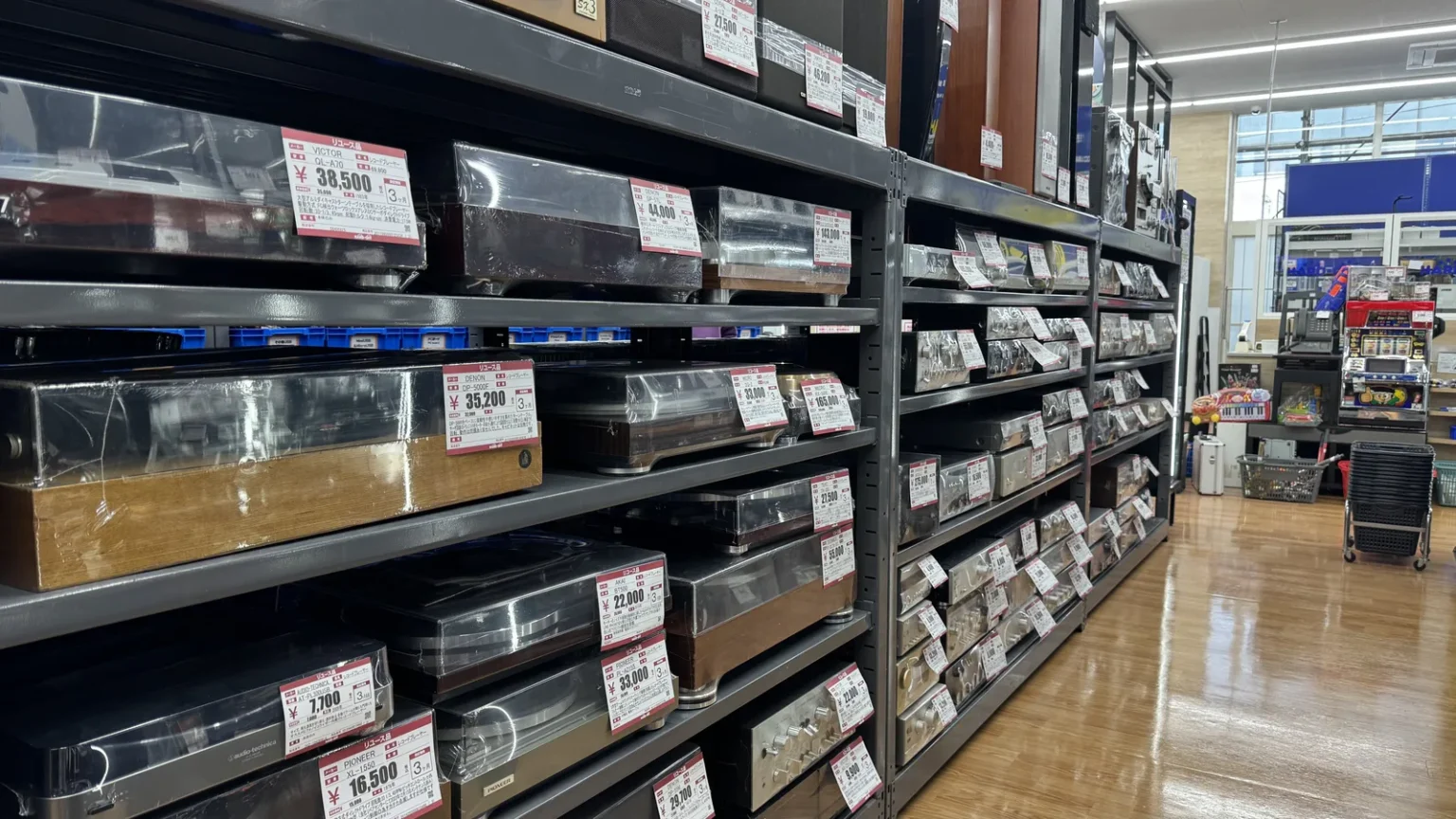 Rows of used turntables and audio equipment on grey metal shelves, with price tags, in a long store aisle