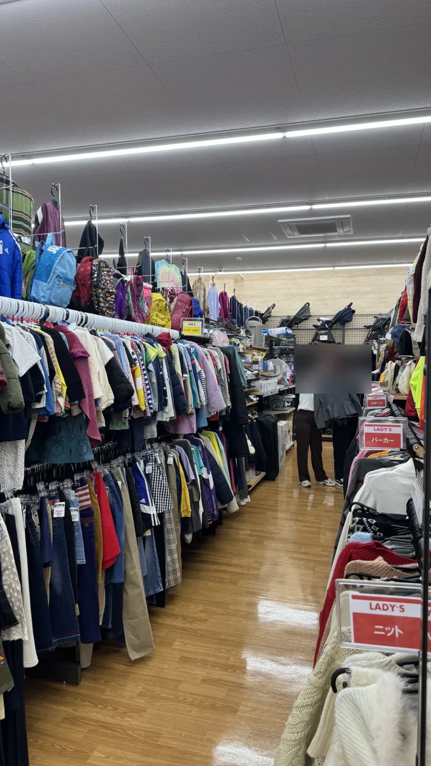 Long racks of used clothing, with shoppers with blurred faces at the end of the aisle
