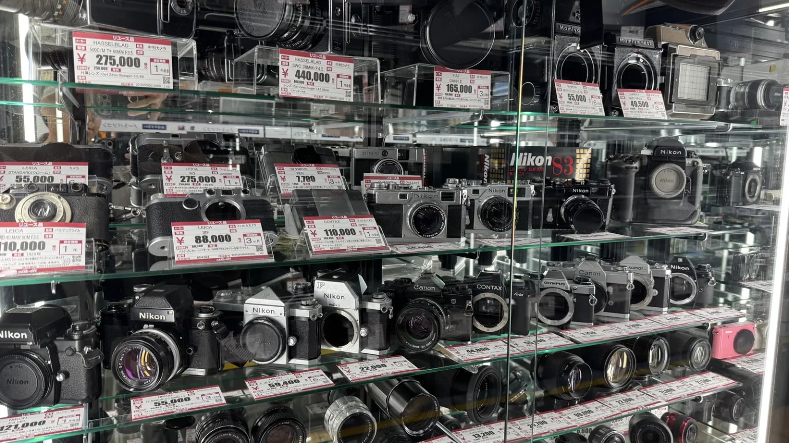 Glass shelves of used film cameras by Leica, Hasselblad, Contax and Olympus and others, with red and white price tags, at Hard Off Kawaguchiko