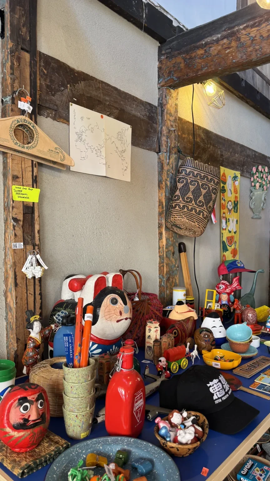 A shop display of red daruma dolls, folk toys, figurines and small baskets on a blue surface against a white wall with dark wooden beams