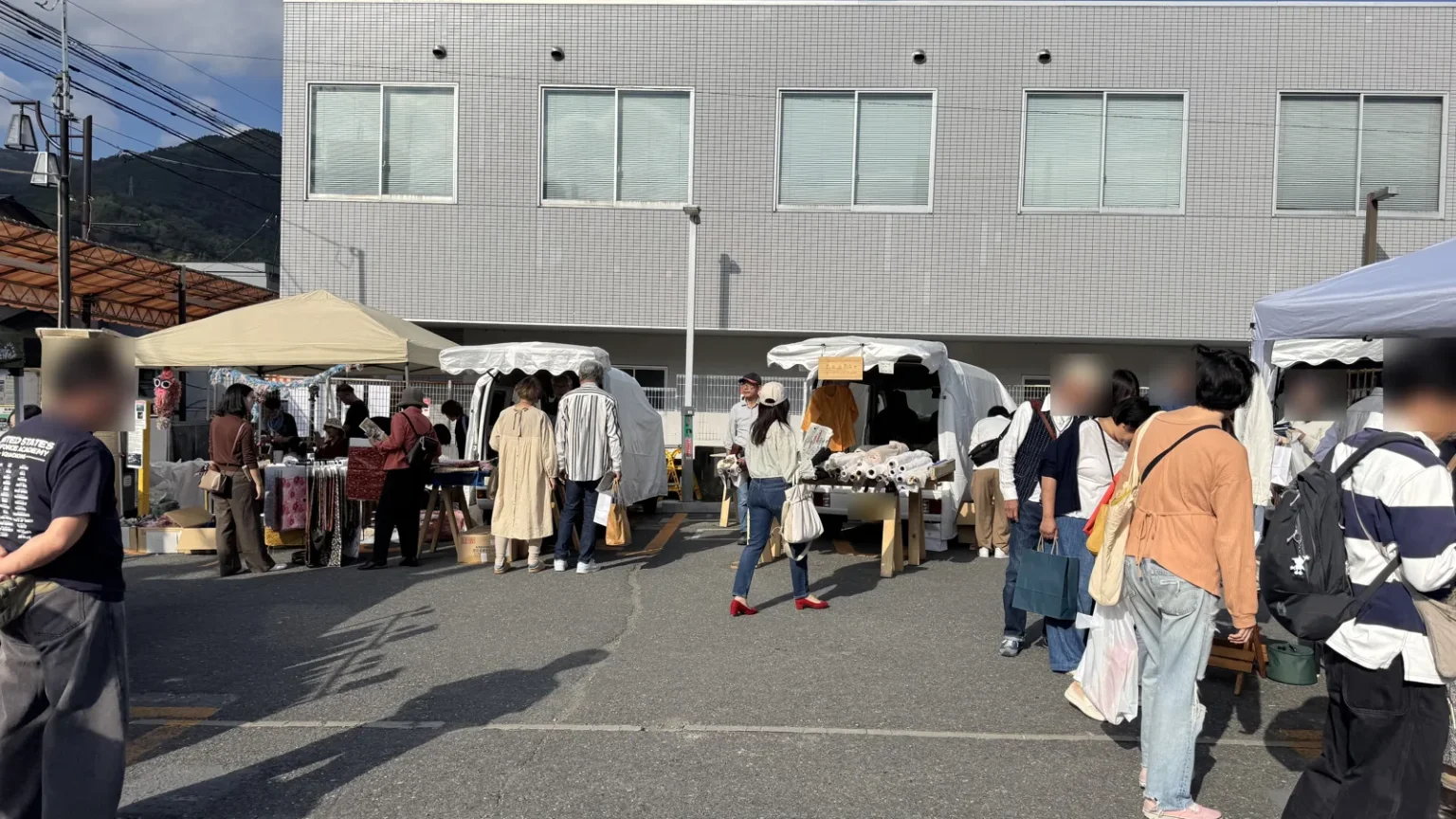 Stalls under white and beige tents and on small trucks in a car park, with shoppers browsing; people close to the camera are blurred