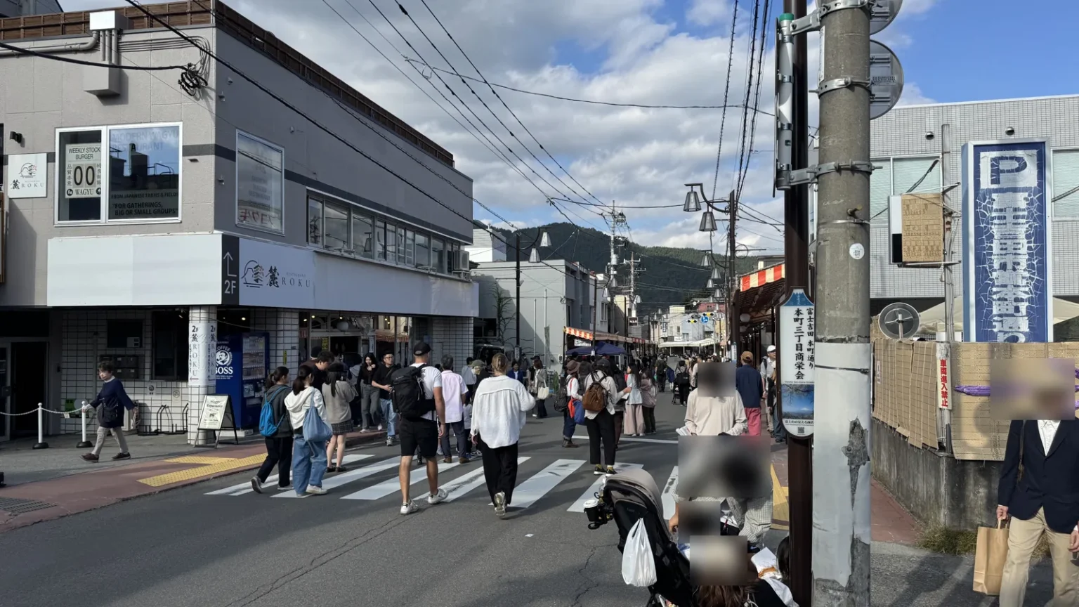 A street crowded with people crossing and walking past shops, by a 本町三丁目商栄会 sign, with wooded hills beyond; people close to the camera are blurred