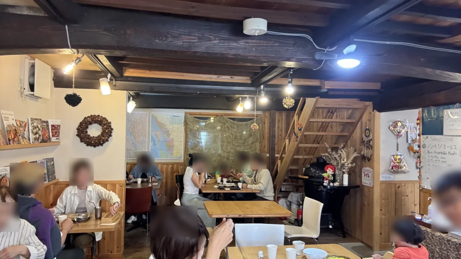 A small restaurant dining room with wooden tables, exposed beams, a wall map of Kerala and a board reading Non-Vegetable Meals; diners close to the camera are blurred