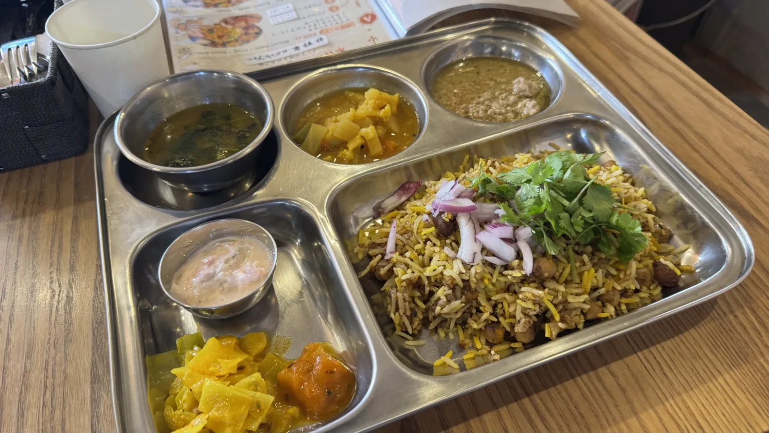 A steel tray with compartments holding rice topped with herbs and red onion, several curries and side dishes, on a wooden table