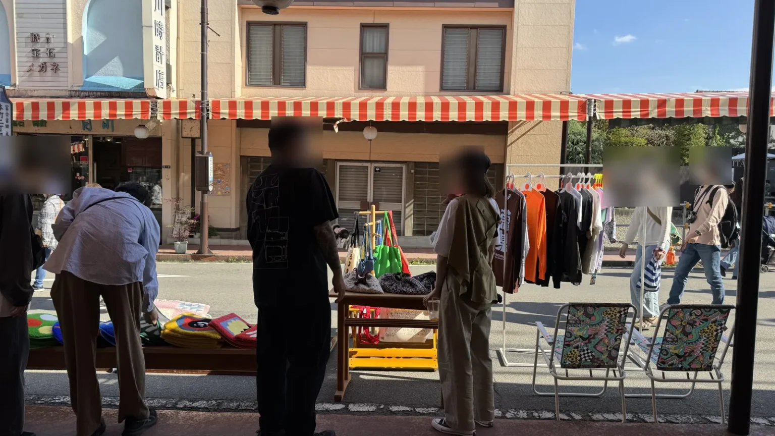 Shoppers at a street stall with a rack of long-sleeve shirts and tees, tote bags on a stand, tufted mats on a table and folding chairs, outside old shopfronts with striped awnings