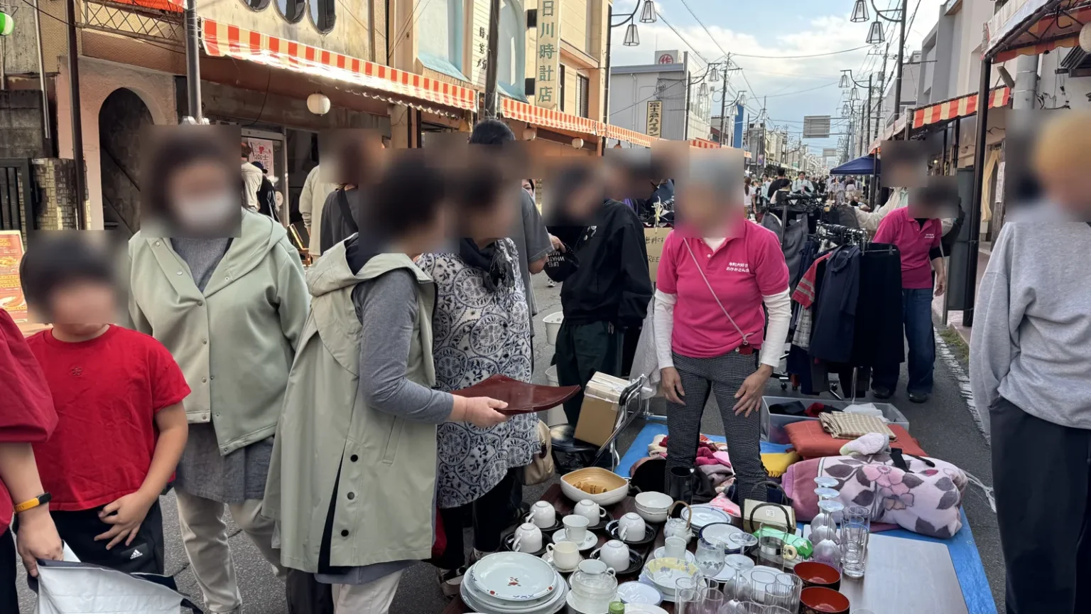 Shoppers around a street stall of cups, plates, glasses and bowls laid out on a low table, with clothing racks behind; people close to the camera are blurred