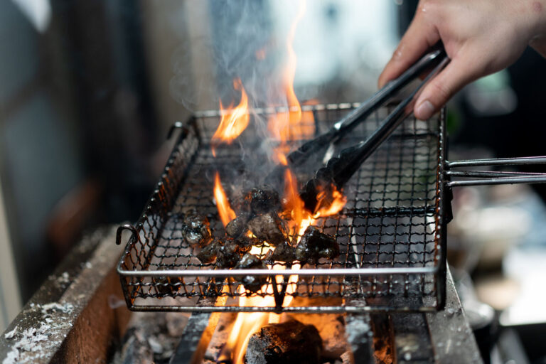 Chicken pieces on a wire grill over open charcoal flame, turned with tongs