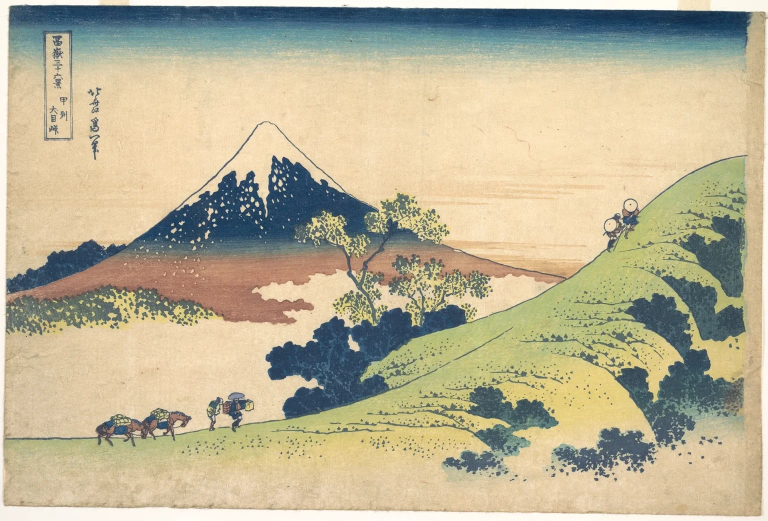Woodblock print: two travellers in round hats climb a bright green hillside at upper right, while two men lead loaded packhorses along the path at lower left. Beyond a sea of pale cloud rises Mount Fuji, white at the top, deep blue on its upper slopes and red-brown at the base.