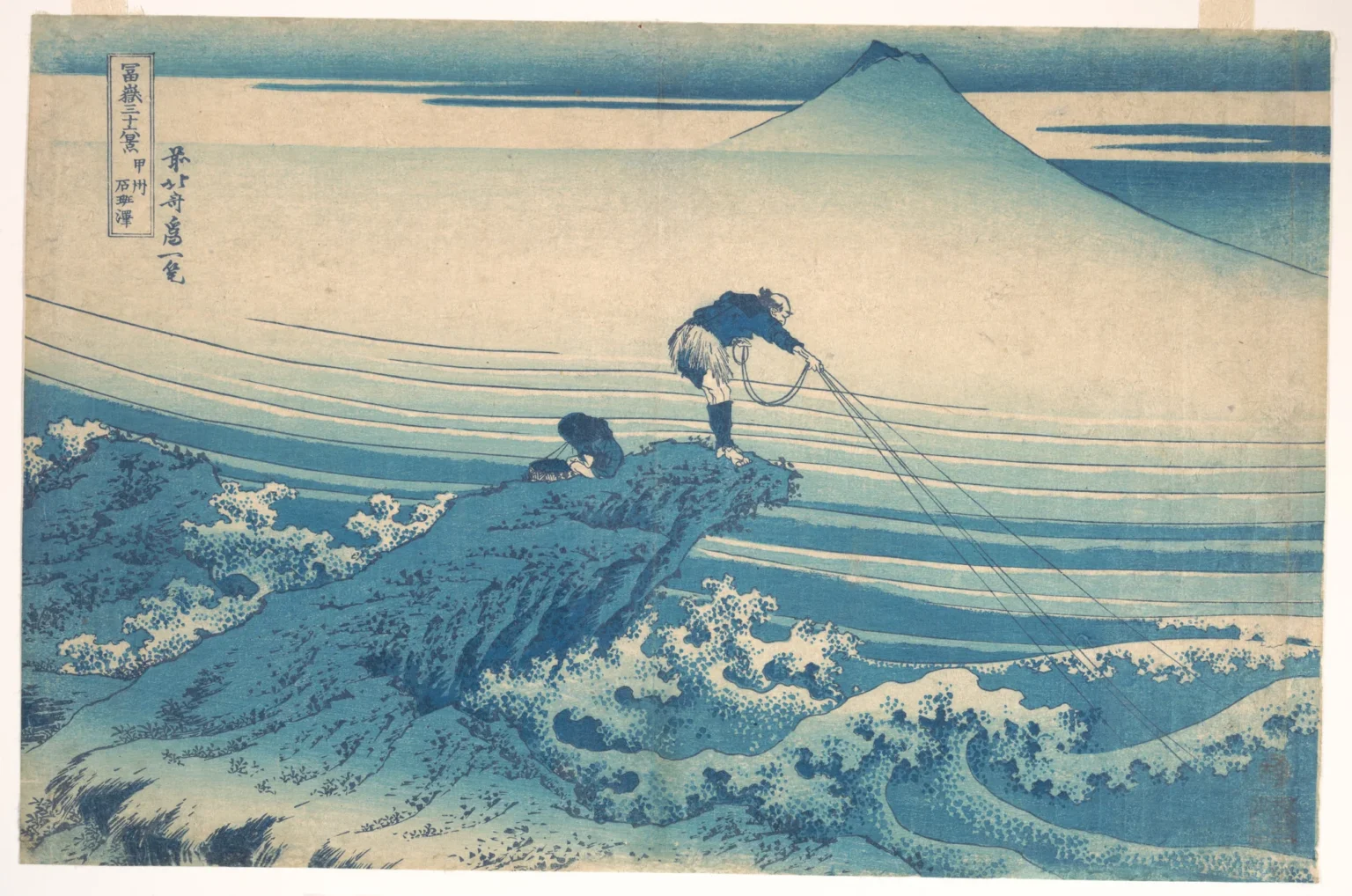Woodblock print in shades of blue: a fisherman stands on a jutting rock above churning, foaming water, leaning forward as the lines of a cast net fan out from his hands, while a boy crouches beside a basket behind him. In the pale distance above the mist rises the outline of Mount Fuji.