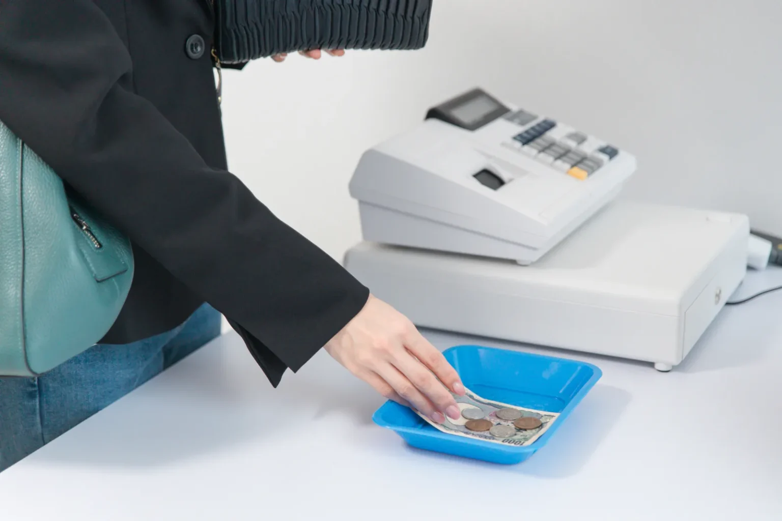 A shopper placing a 1,000 yen note and coins in a blue cash tray beside a white cash register