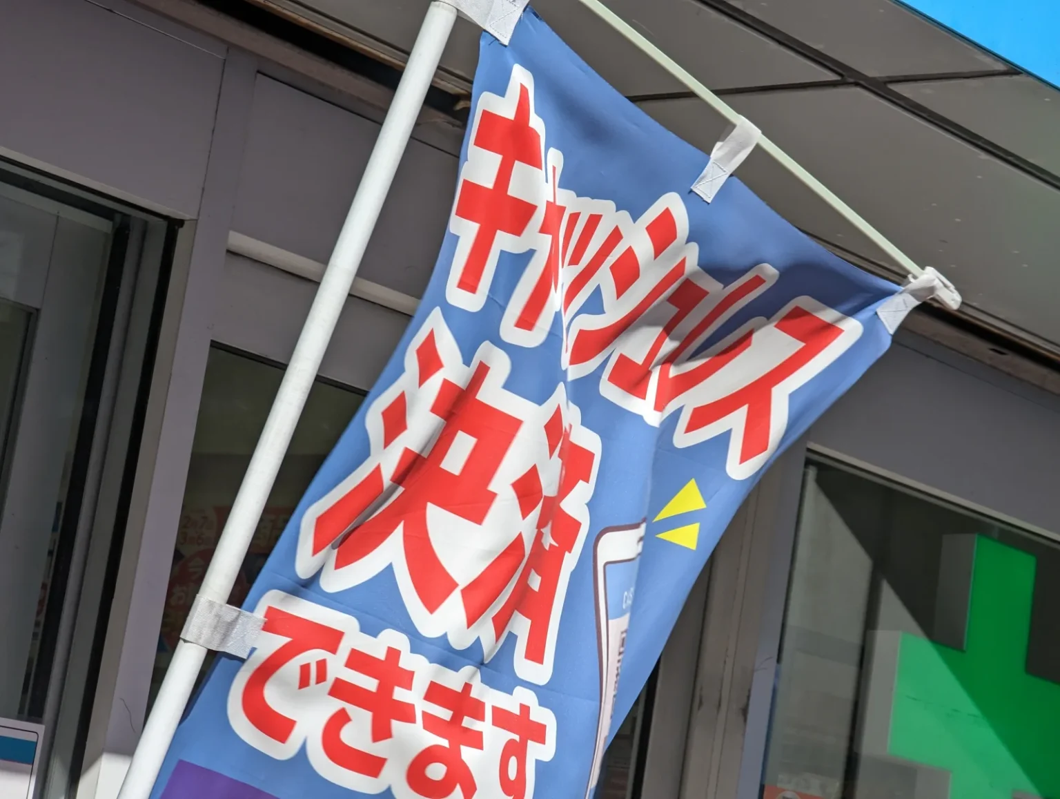 A blue banner with large red Japanese text reading “cashless payment accepted” on a pole outside a shop front