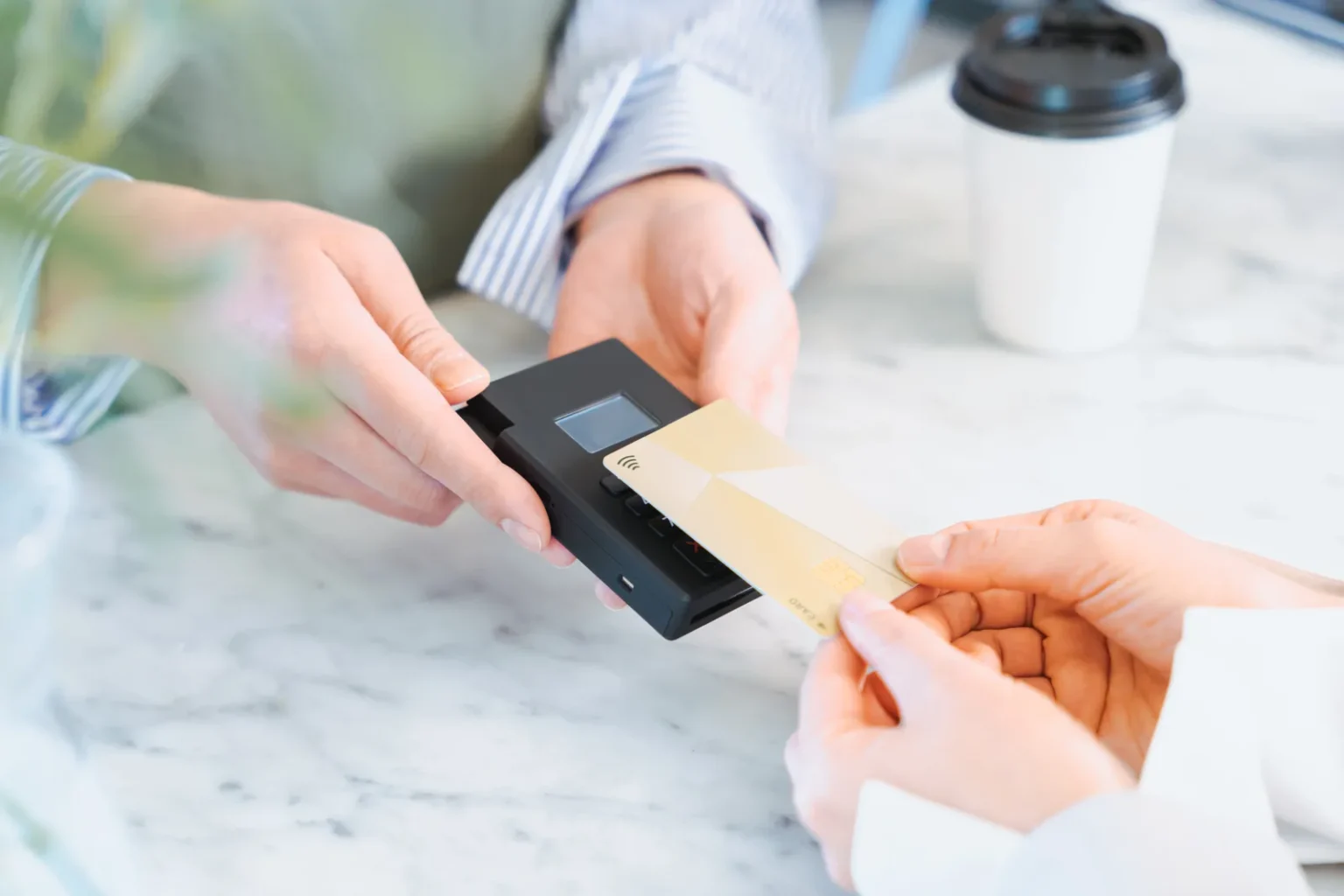 A gold payment card held against a small black card reader on a marble café counter, next to a takeaway coffee cup