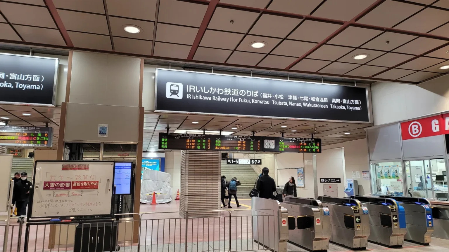 A row of automatic ticket gates under an IR Ishikawa Railway platform sign at Kanazawa Station, with departure boards above and passengers walking through