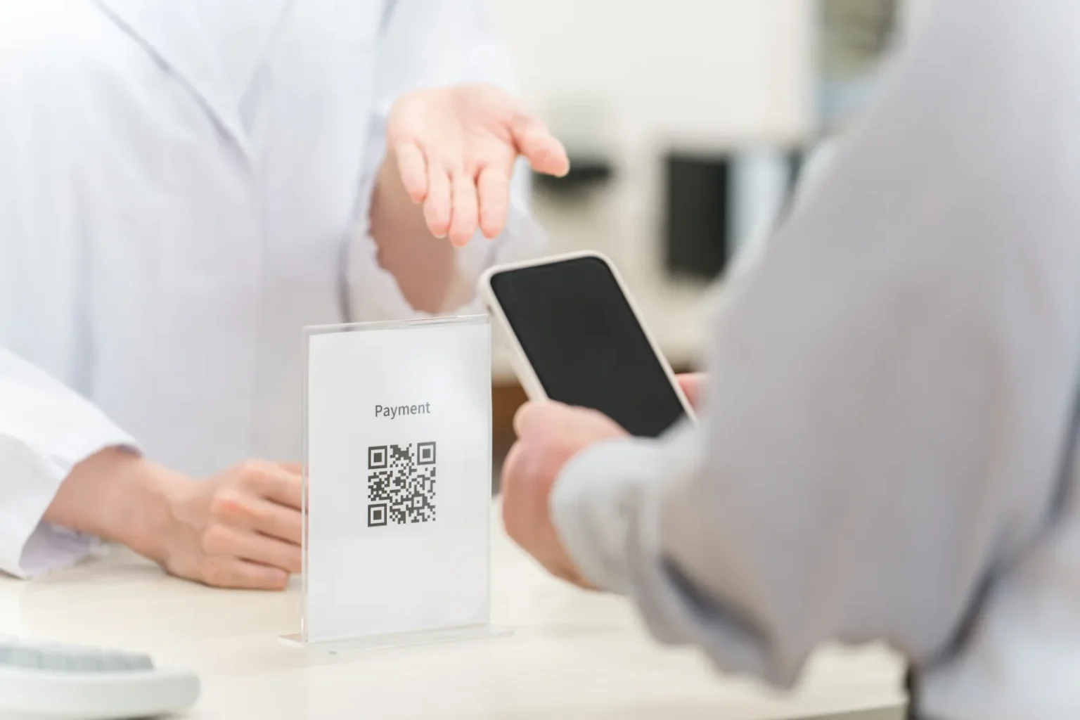 A clear acrylic stand with a QR code and the word Payment on a counter, a customer holding a smartphone and a staff member gesturing towards the stand