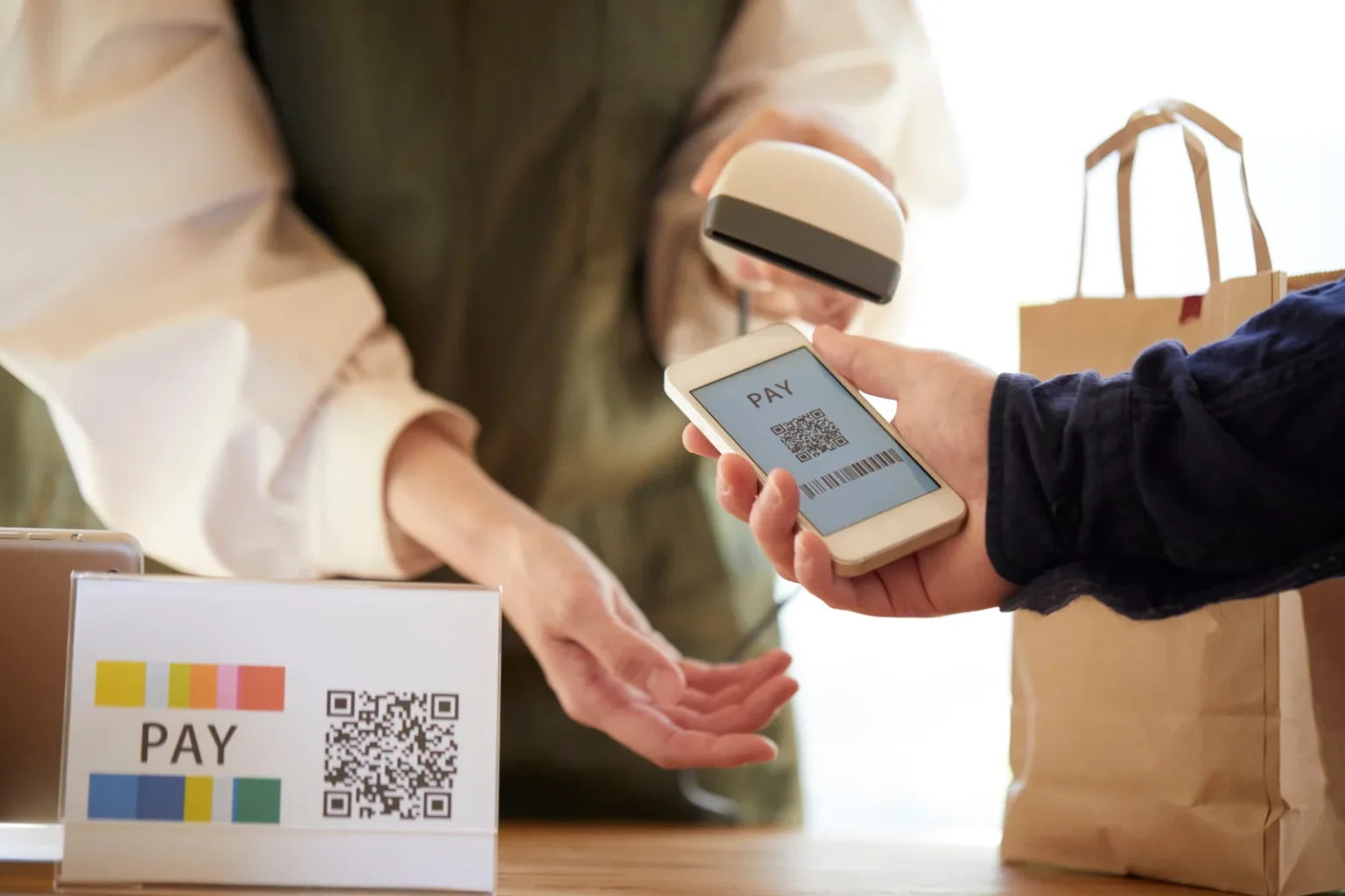 A shop worker scanning a code on a customer’s smartphone with a handheld scanner, beside a counter sign with a QR code and the word PAY